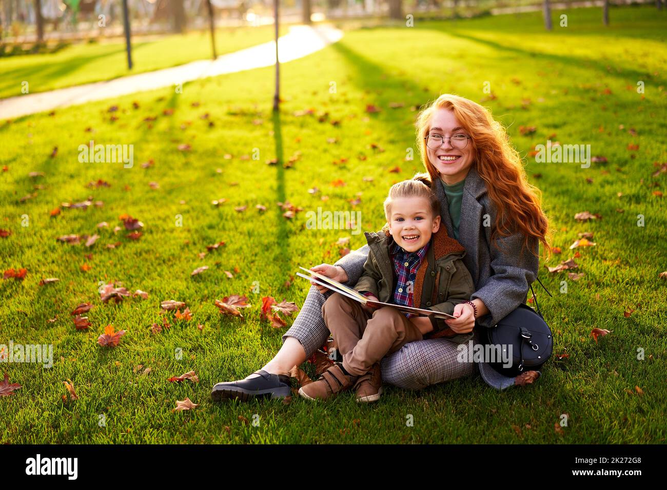 Rotschopf Frau liest Buch mit kleinen Jungen auf Gras im Stadtpark sitzen. Die junge Mutter unterrichtet ihren Sohn mit der ersten Klasse und macht Hausaufgaben am sonnigen Herbsttag Stockfoto