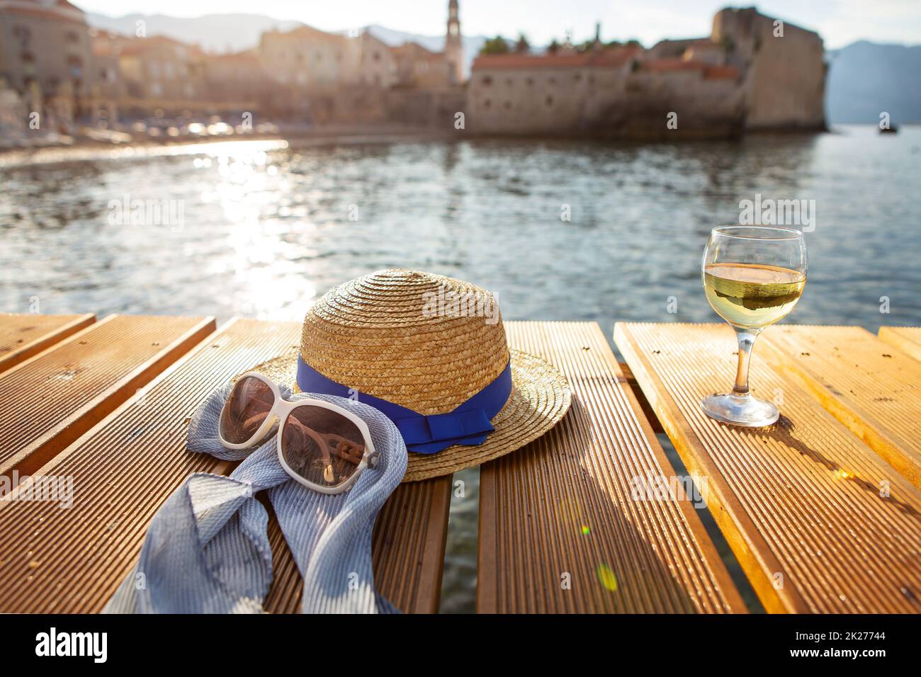 Ein Strohhut, Gläser und ein Glas Wein stehen auf einem Pier nahe dem Meer, aus nächster Nähe. Schönes Licht von der Sonne. Platz für Text. Stockfoto