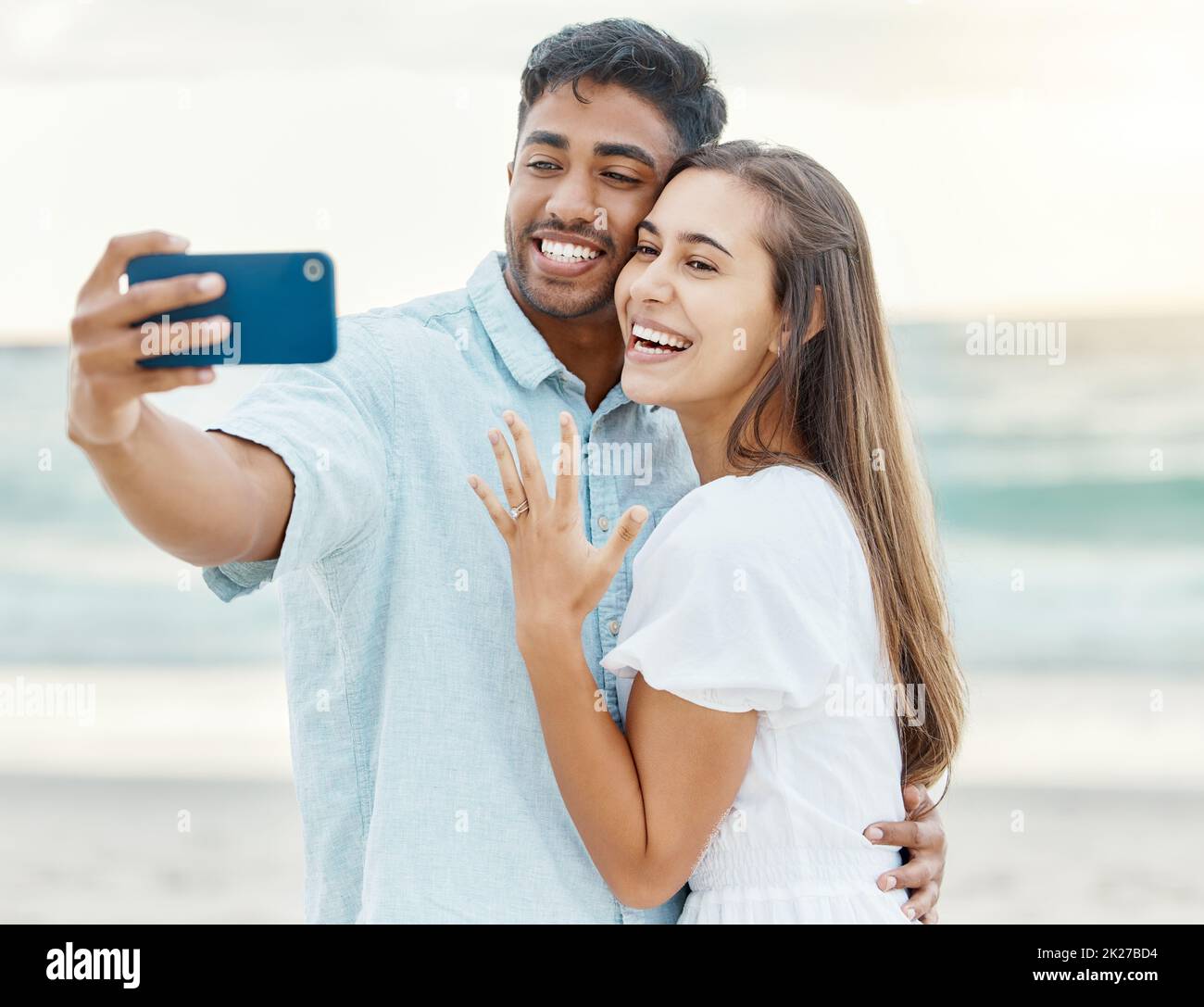 Liebe, Strand und Paar nach ihrer Verlobung ein Selfie auf einem Telefon während der Sommerferien. Glückliche Frau, die nach einem Vorschlag ihren Ring ablegt Stockfoto