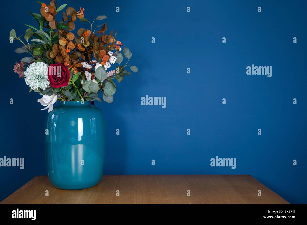 Blaue Vase auf Holztisch mit künstlichem Blumenstrauß aus Kunststoff an der blauen Wand, moderne, stilvolle Dekoration im Wohnzimmer mit Kopierbereich. Dekorationskonzept Stockfoto