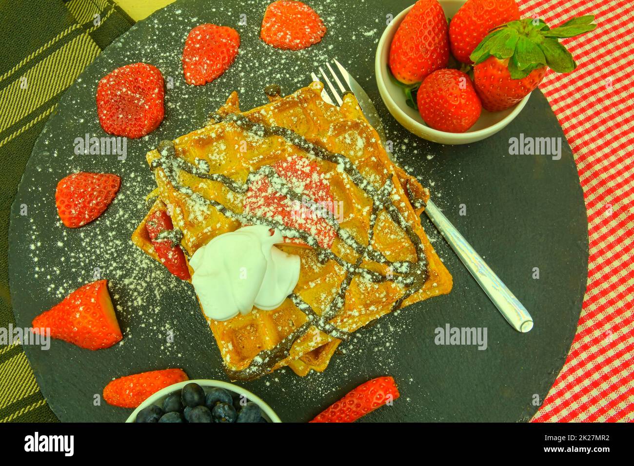 Quadratische belgische Waffeln mit Beeren und Puderzucker auf einem schwarzen Teller. Leckere süße, zuckerhaltige Waffeln. Geschmolzene Schokolade und Schlagsahne auf Waffeln. Draufsicht Stockfoto