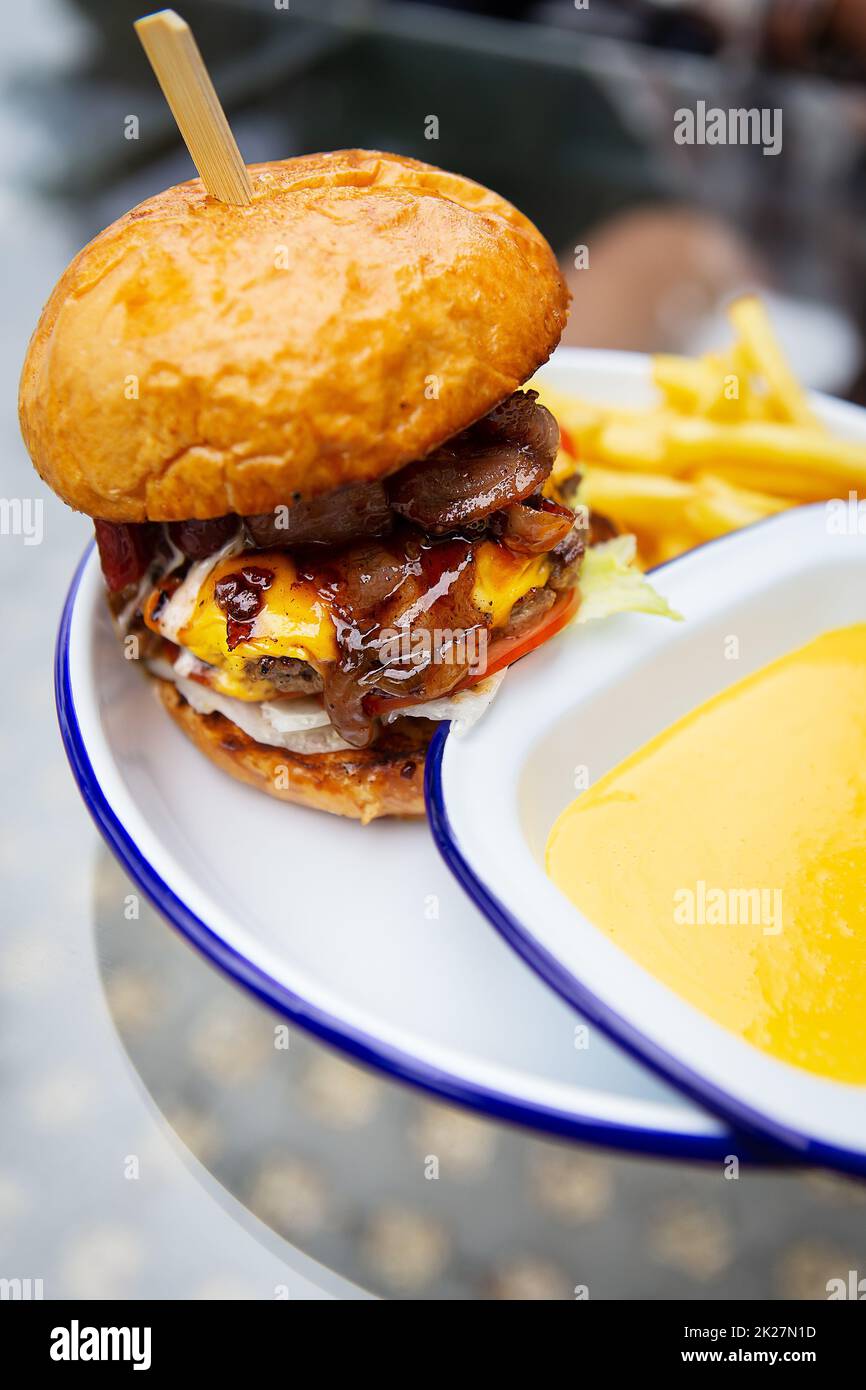 Das Konzept von Fast Food und Essen zum Mitnehmen. Ein saftiger Hamburger liegt neben pommes frites auf einem Metallteller zusammen mit Cheddar Cheese Sauce im Hintergrund. Stockfoto