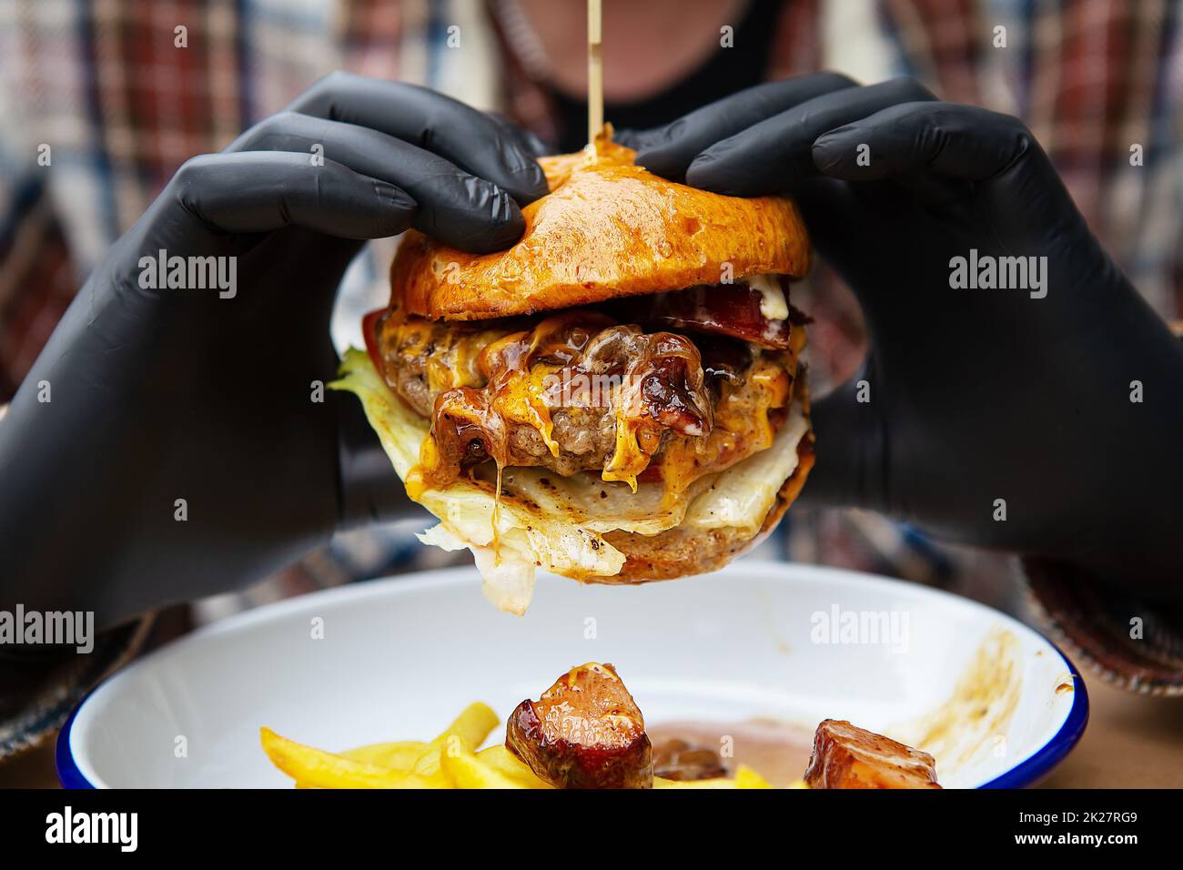Das Konzept von Fast Food und Essen zum Mitnehmen. Ein Mann in schwarzen Latexhandschuhen hält einen saftigen Hamburger in den Händen, liegt neben pommes Frites auf einem Metallteller zusammen mit Käsesauce. Stockfoto