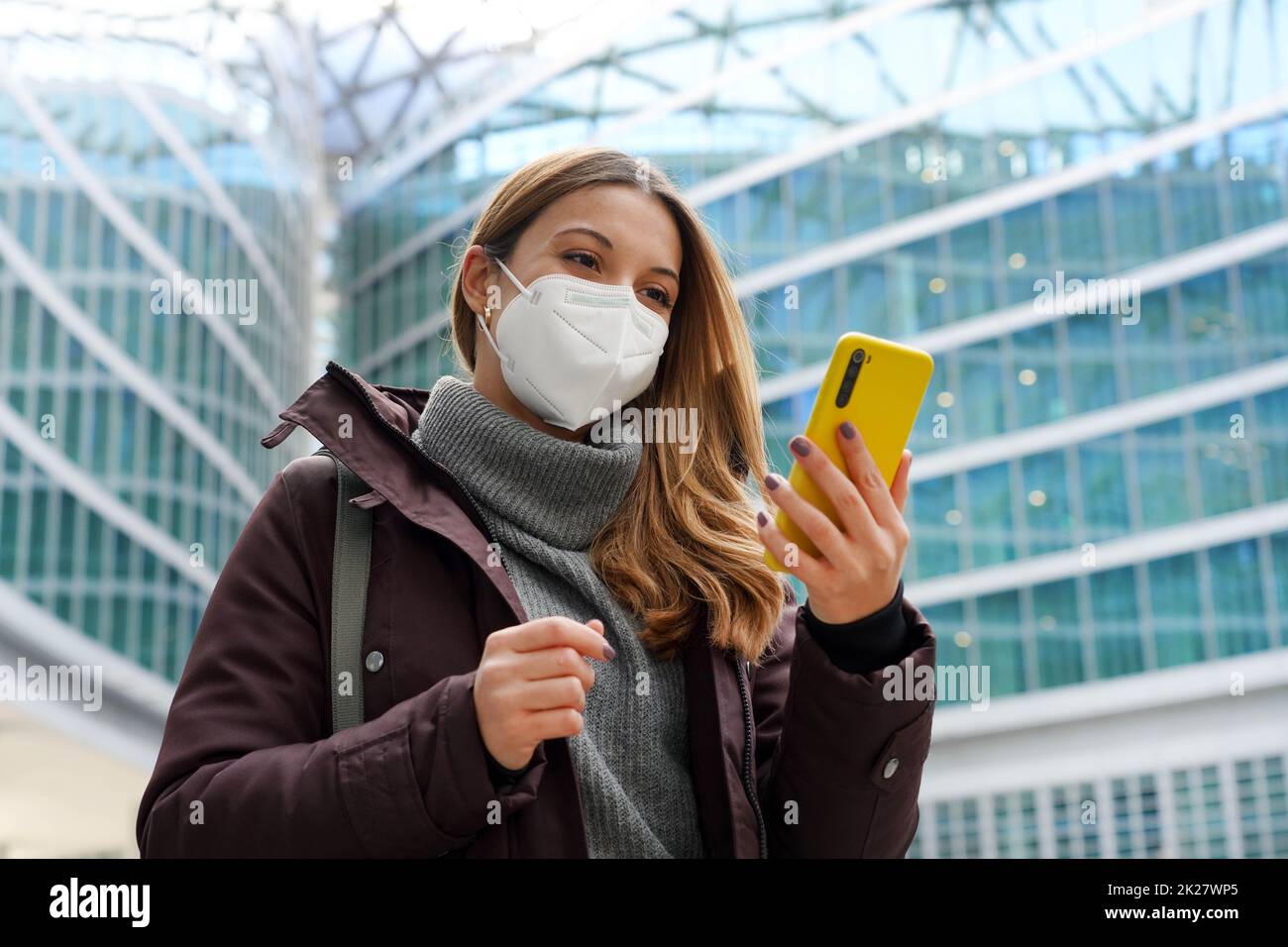 Geschäftsfrau, die das Gesicht mit einer Schutzmaske bedeckt, die das Mobiltelefon mit modernem Stadthintergrund hält. Technologie, Wachstum, Gesundheitskonzept. Stockfoto