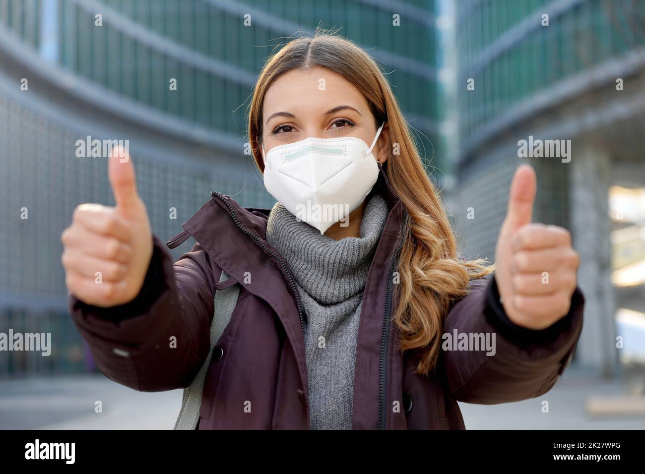 Porträt des optimistischen Mädchen trägt schützende Gesichtsmaske zeigt Daumen nach oben in der modernen Stadt Straße und Blick auf die Kamera Stockfoto