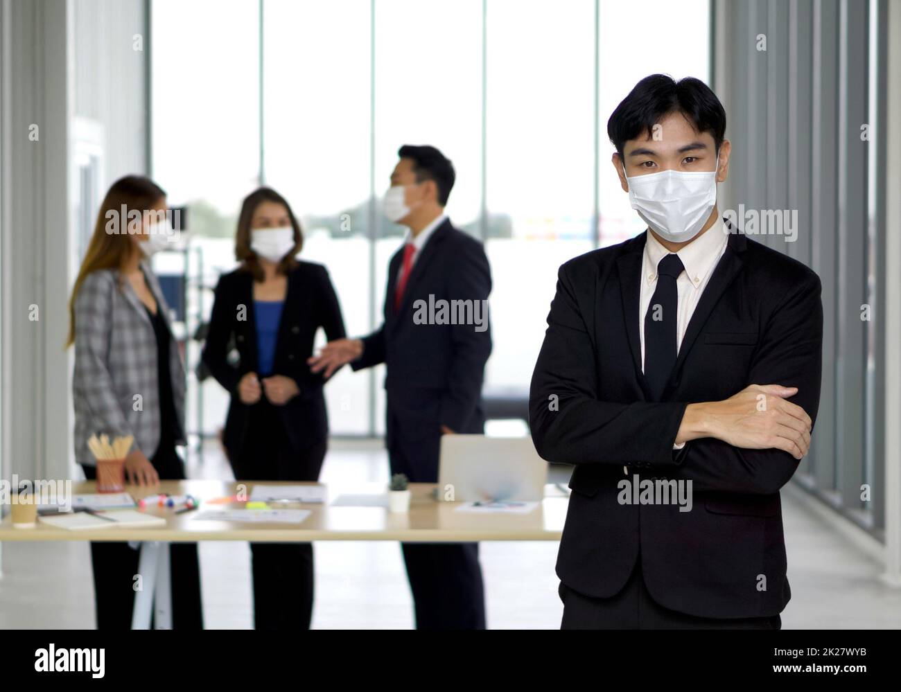 Ein junger asiatischer Geschäftsmann im schwarzen Anzug faltete seine Arme selbstbewusst über die Brust. Das Team bereitet sich im Hintergrund auf das Meeting vor. Jeder trägt eine Gesichtsmaske. Stockfoto