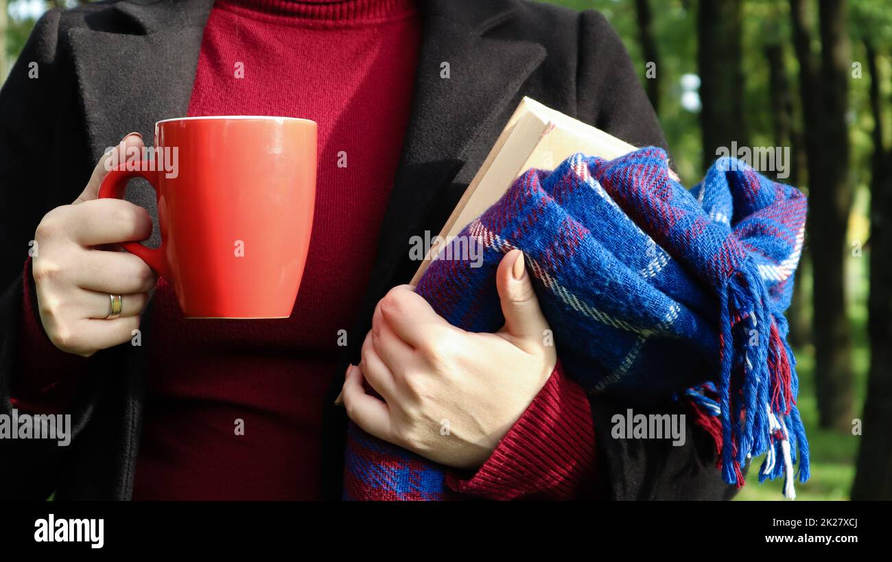 Eine rote Tasse Kaffee und ein Buch mit einer blauen karierten Wolldecke oder in den Händen einer Frau, die im Park einen Pullover und einen schwarzen Mantel trägt. Warmes und sonniges Wetter. Weiche, gemütliche Fotografie Stockfoto