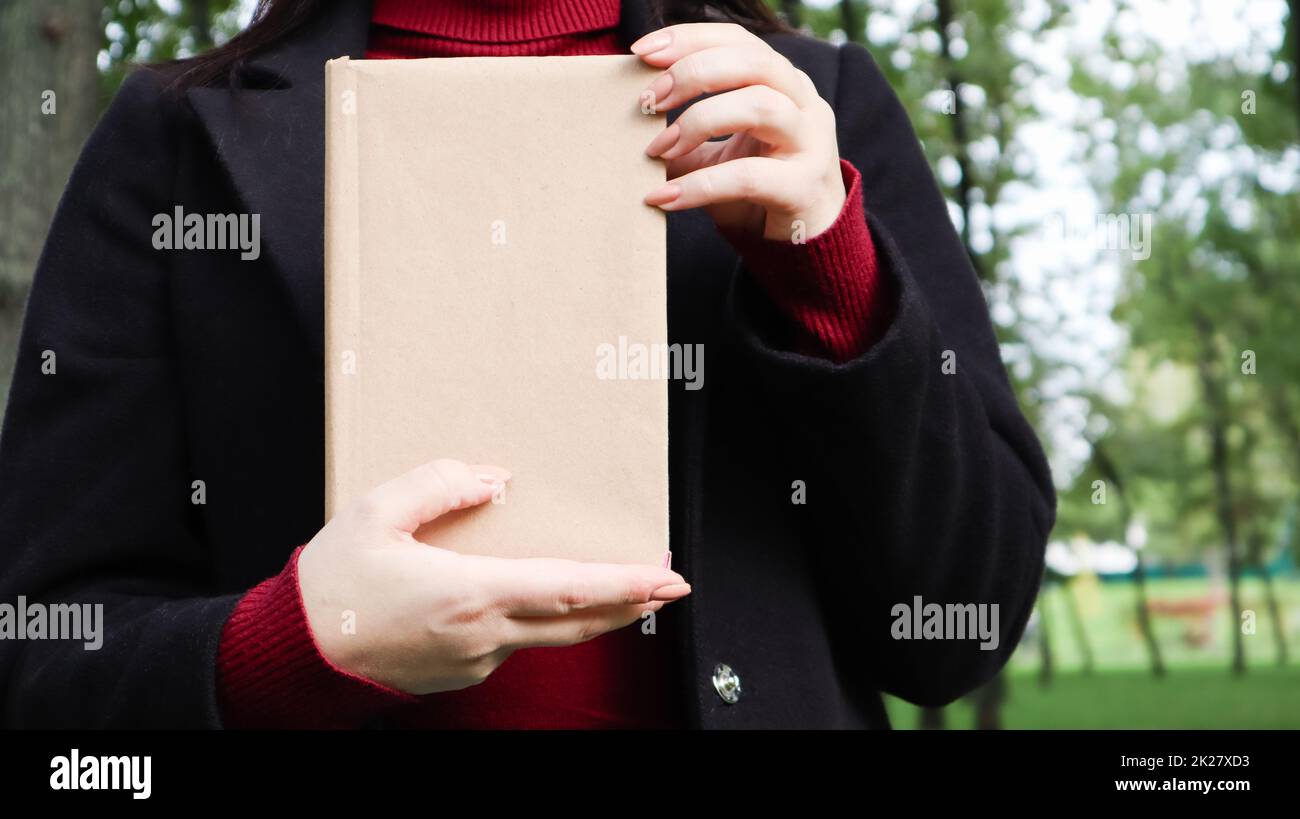 Nahaufnahme einer Frau in schwarzem Wollmantel und burgunderfarbenem Pullover mit einem Buch mit leerem Einband im Park. Freier Platz für das Hintergrundlayout Ihres Buchlesekonzepts. Stockfoto
