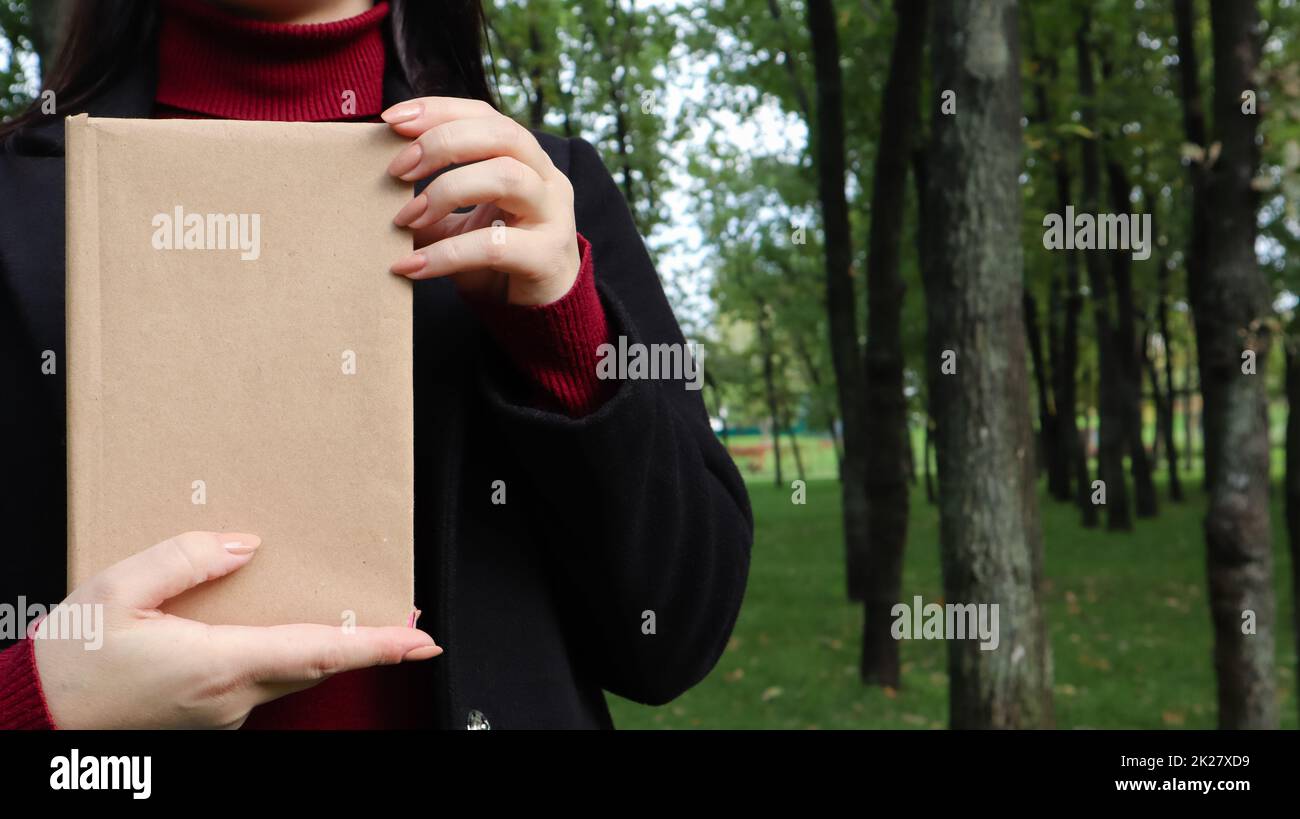 Nahaufnahme einer Frau in schwarzem Wollmantel und burgunderfarbenem Pullover mit einem Buch mit leerem Einband im Park. Freier Platz für das Hintergrundlayout Ihres Buchlesekonzepts. Stockfoto