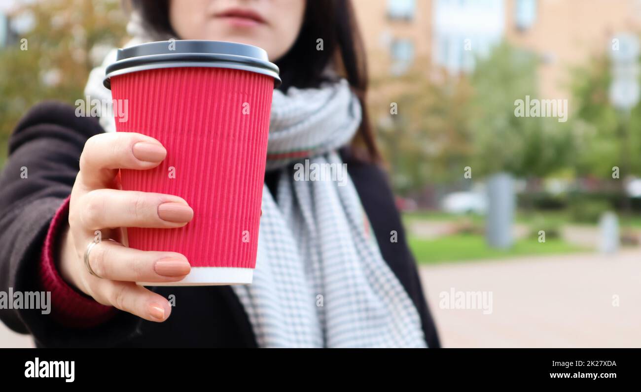 Eine junge, stilvolle Frau in Mantel und Schal trinkt morgens heißen Kaffee in einem roten, umweltfreundlichen Papierglas draußen in einem Herbstpark. Nahaufnahme einer jungen Frau mit einer Kaffeetasse zum Mitnehmen, geringe Schärfentiefe Stockfoto