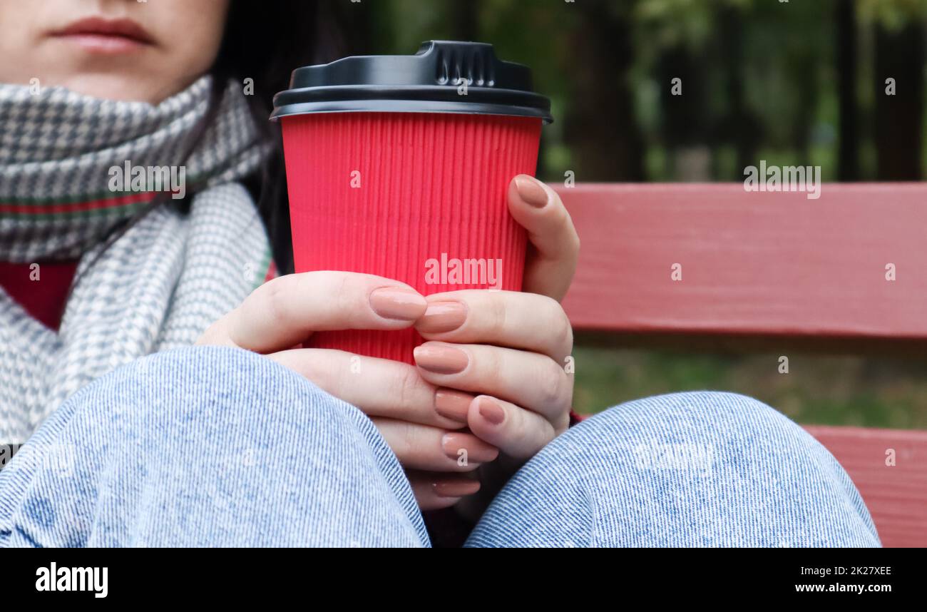 Eine junge, stilvolle Frau in Mantel und Schal trinkt morgens heißen Kaffee in einem roten, umweltfreundlichen Papierglas draußen in einem Herbstpark. Nahaufnahme einer jungen Frau mit einer Kaffeetasse zum Mitnehmen, geringe Schärfentiefe Stockfoto