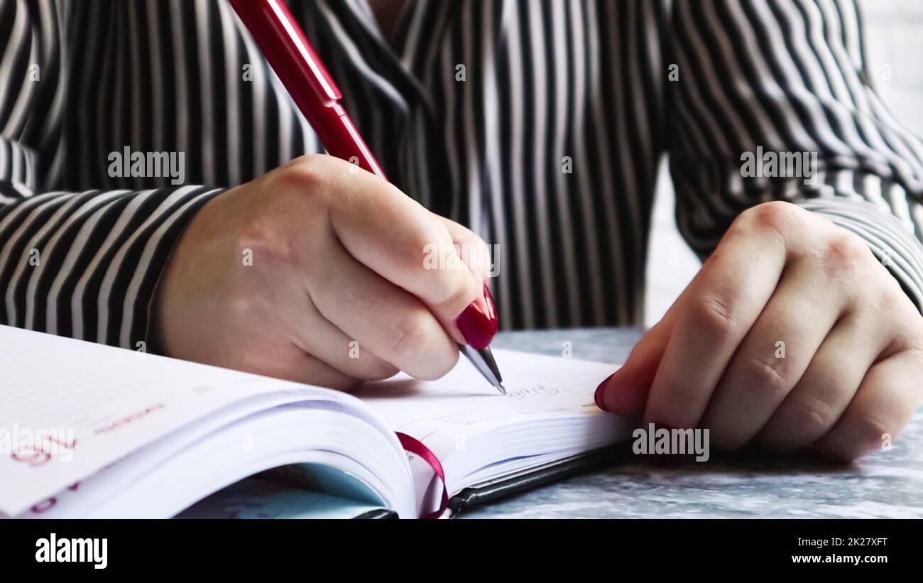 Seitenansicht der Hände einer Frau mit roten Nägeln, mit einem roten Stift, schreibt etwas auf ein leeres Blatt mit Tagebüchern, das auf einem grauen Tisch liegt. Ein Mädchen macht Notizen in einem schwarzen Notizbuch Stockfoto