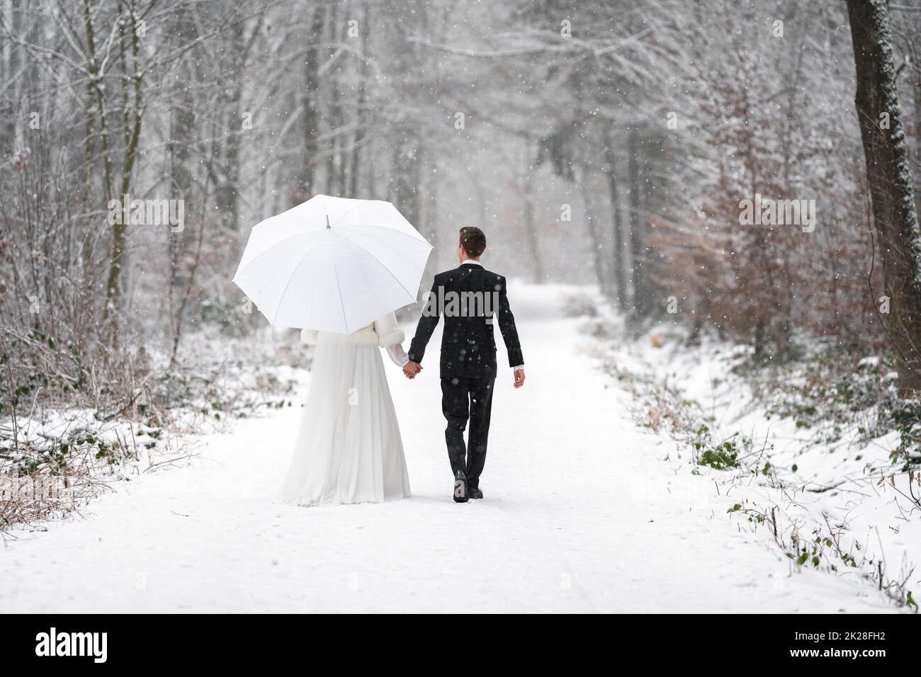 Brautpaar in Hochzeitskleider im winterverschneiten Wald auf einem Spaziergang mit einem Regenschirm Stockfoto