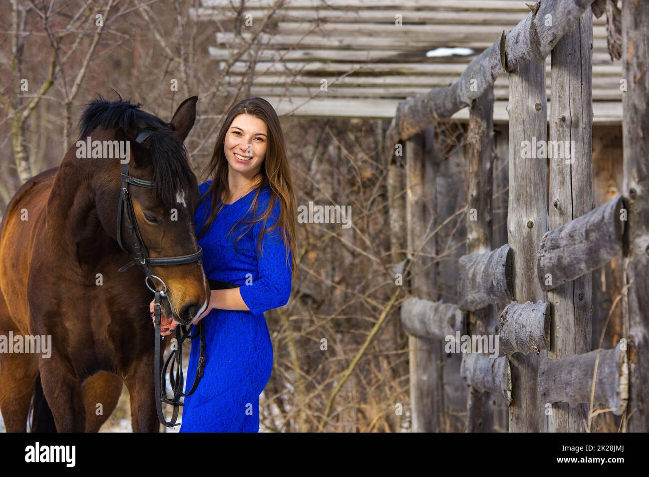 Ein schönes junges Mädchen in einem blauen Kleid umarmt ein Pferd vor dem Hintergrund eines alten Zauns und eines Winterwaldes Stockfoto