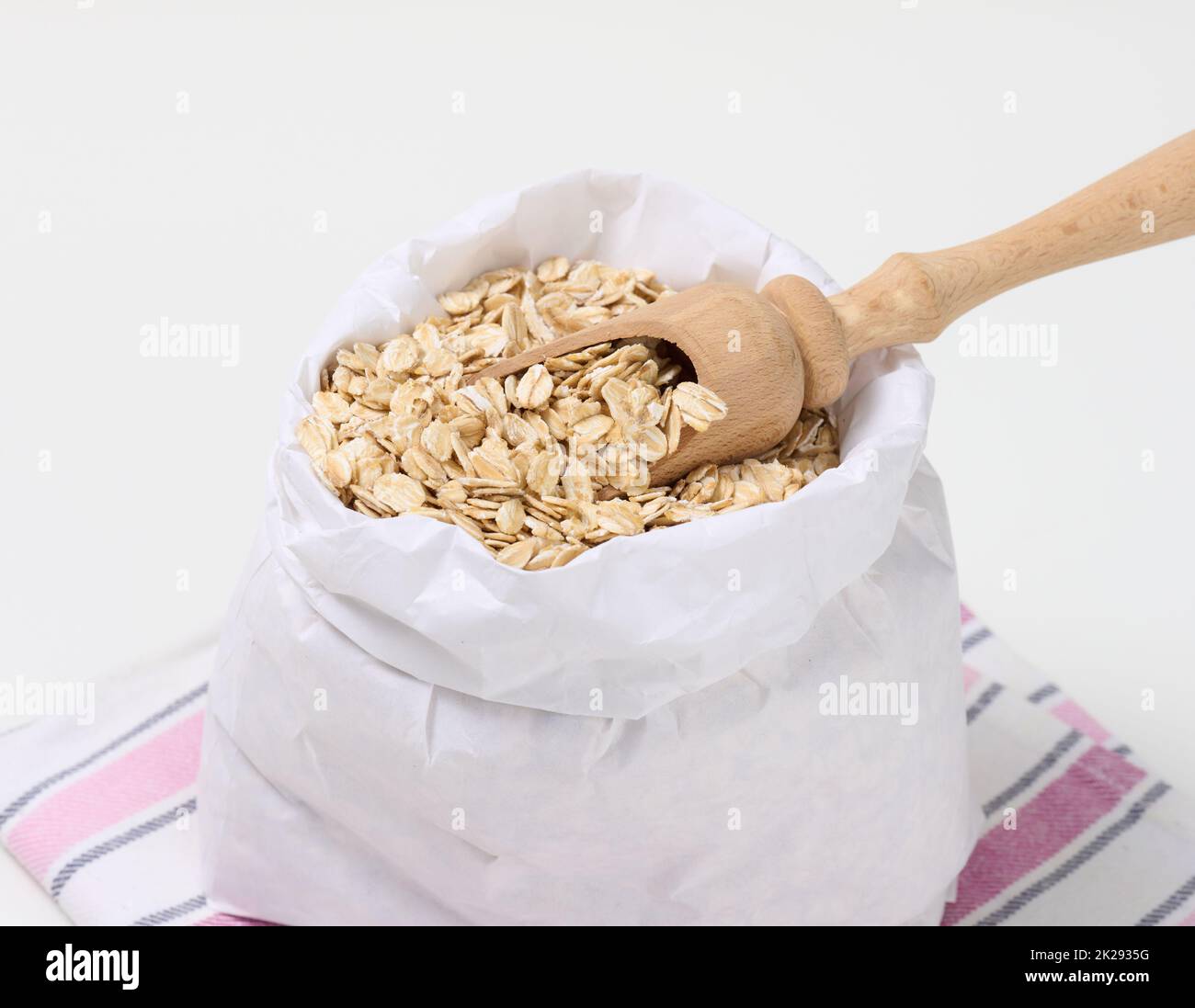 Roher Haferbrei in einem weißen Papierbeutel und ein Holzlöffel auf einem weißen Tisch, Frühstücksbrei Stockfoto