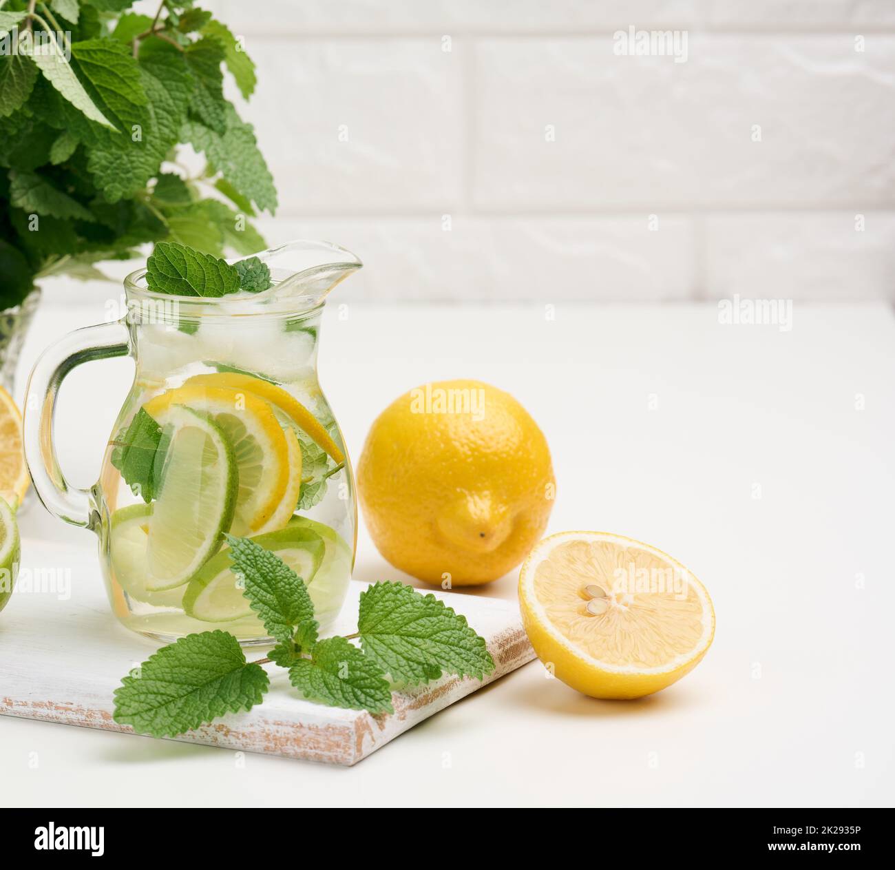 Transparente Glasdekanter mit Scheiben von Zitronen-, Limetten- und Minzblättern auf einem weißen Tisch, entgiften. Hinter den Zutaten für das Getränk Stockfoto