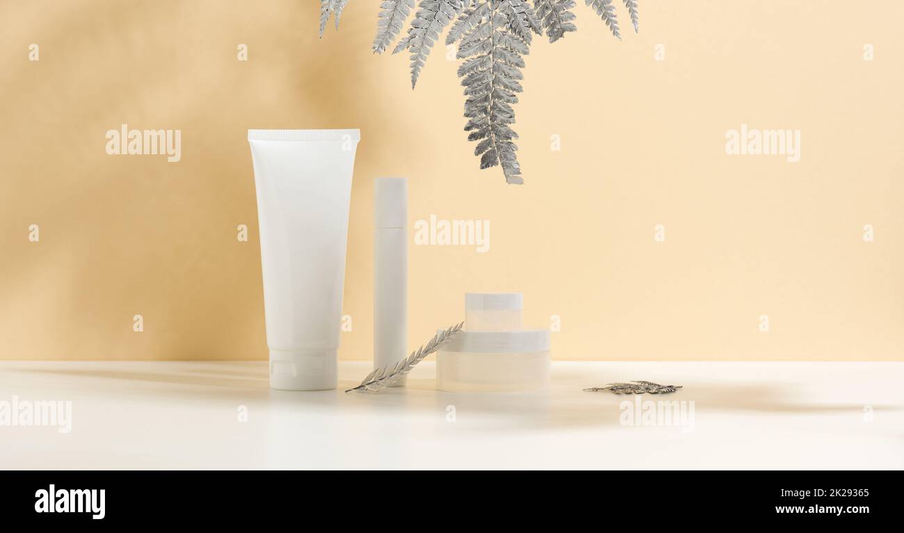 Weiße Tube für Kosmetik, ein Glas Creme und silberne Blätter auf einem weißen Tisch. Kosmetik auf beigefarbenem Hintergrund. Cremeflasche, Lotion, Reinigungsmittel, Shampoo für die Hautpflege Stockfoto