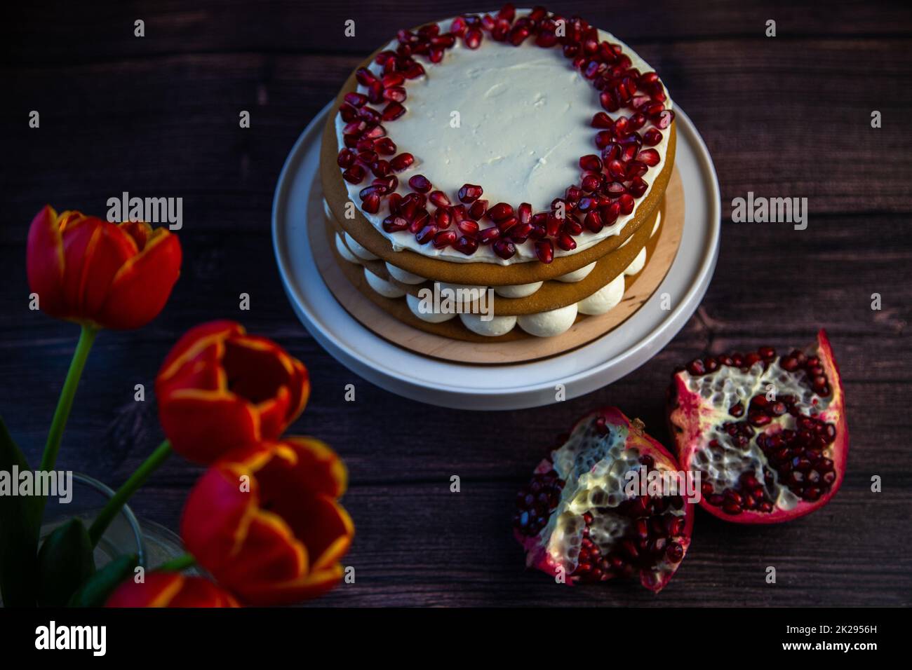 Ein dreischichtiger Kuchen aus Kuchen und weißer Sahne, dekoriert mit Granatapfel oben in einem Kreis, steht auf dunklem Hintergrund neben Tulpen und Granatäpfeln. Stockfoto