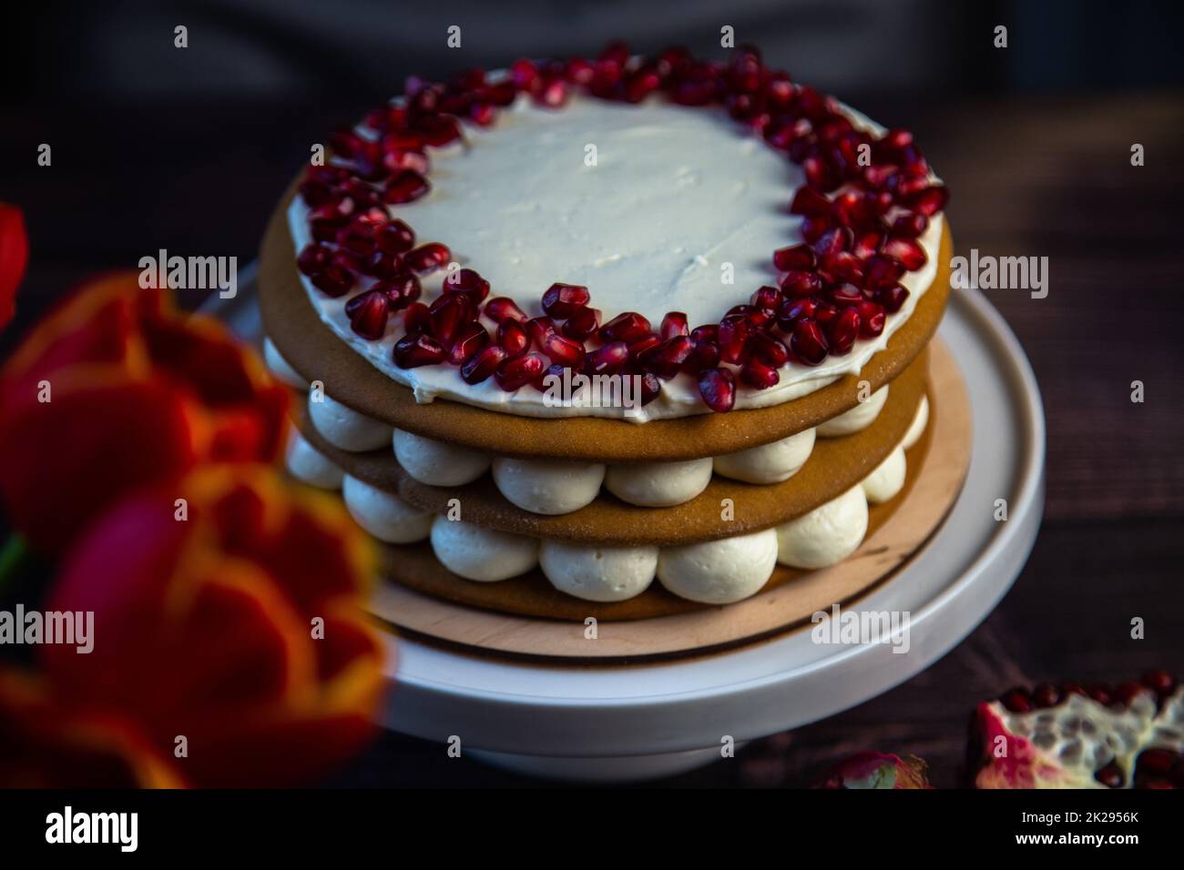 Ein dreischichtiger Kuchen aus Kuchen und weißer Sahne, dekoriert mit Granatapfel oben in einem Kreis, steht auf dunklem Hintergrund neben Tulpen und Granatäpfeln. Stockfoto