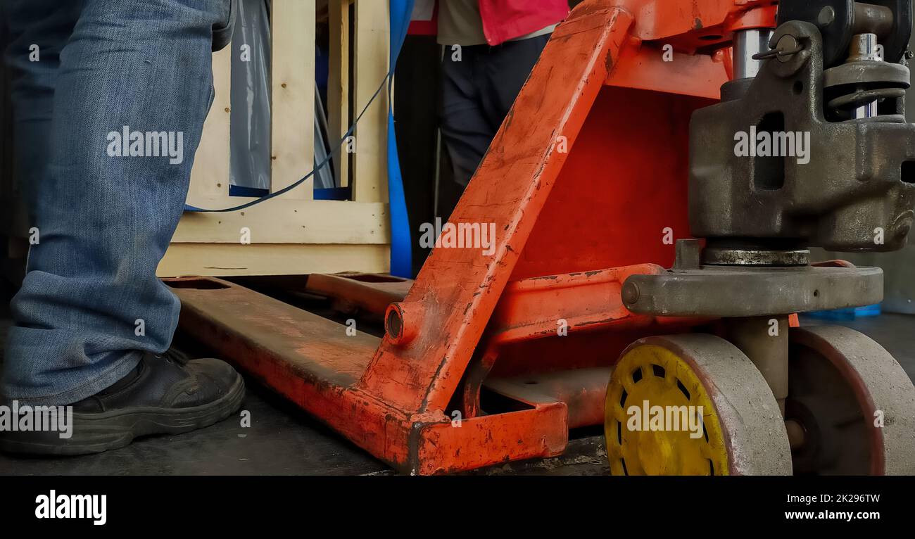 Gabelstapler, der Holzkisten der Fabrikmaschine im Lager transportiert. Arbeiter mit Sicherheitsschuhen, der mit Handgabelhubwagen arbeitet. Güterverkehr. Lager- und Logistikgeschäft. Orangefarbener Niederhubwagen. Stockfoto