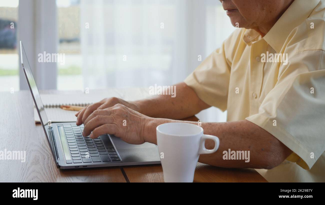 Ein älterer Mann trägt eine Brille, sitzt auf einem Stuhl und arbeitet an einem Laptop Stockfoto