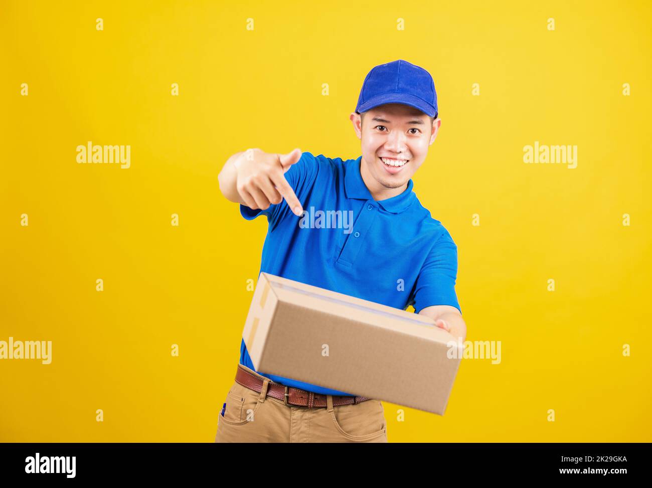 Lieferung glücklicher Mann logistisch stehend lächelt er trägt ein blaues T-Shirt und eine Kappe Uniform hält Paketkiste mit dem Finger auf die Kiste zeigt Stockfoto