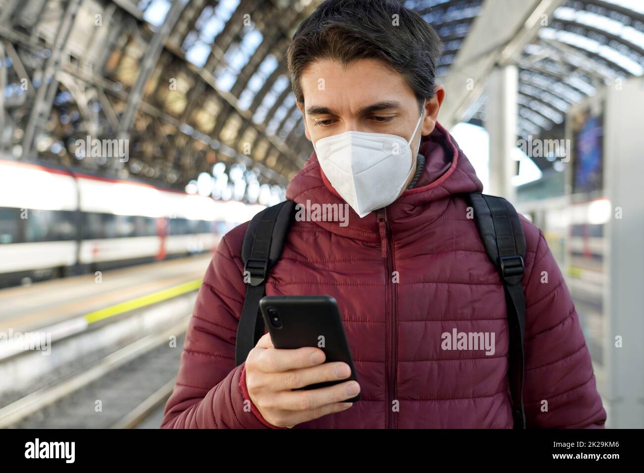 Junger Reisender Mann mit KN95 FFP2 Gesichtsmaske am Bahnhof. Junger kaukasischer Mann, der im Bahnhof ein Smartphone benutzt. Stockfoto
