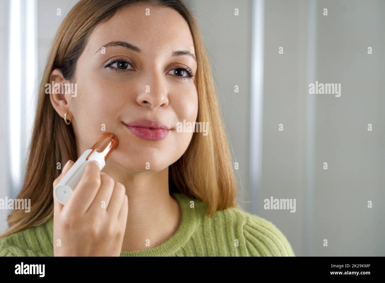 Elektrischer Epilierer für das Gesicht. Nahaufnahme der schönen Mädchen sieht sich in den Spiegel mit Haarentfernung zu Hause. Gesichtspflege. Stockfoto