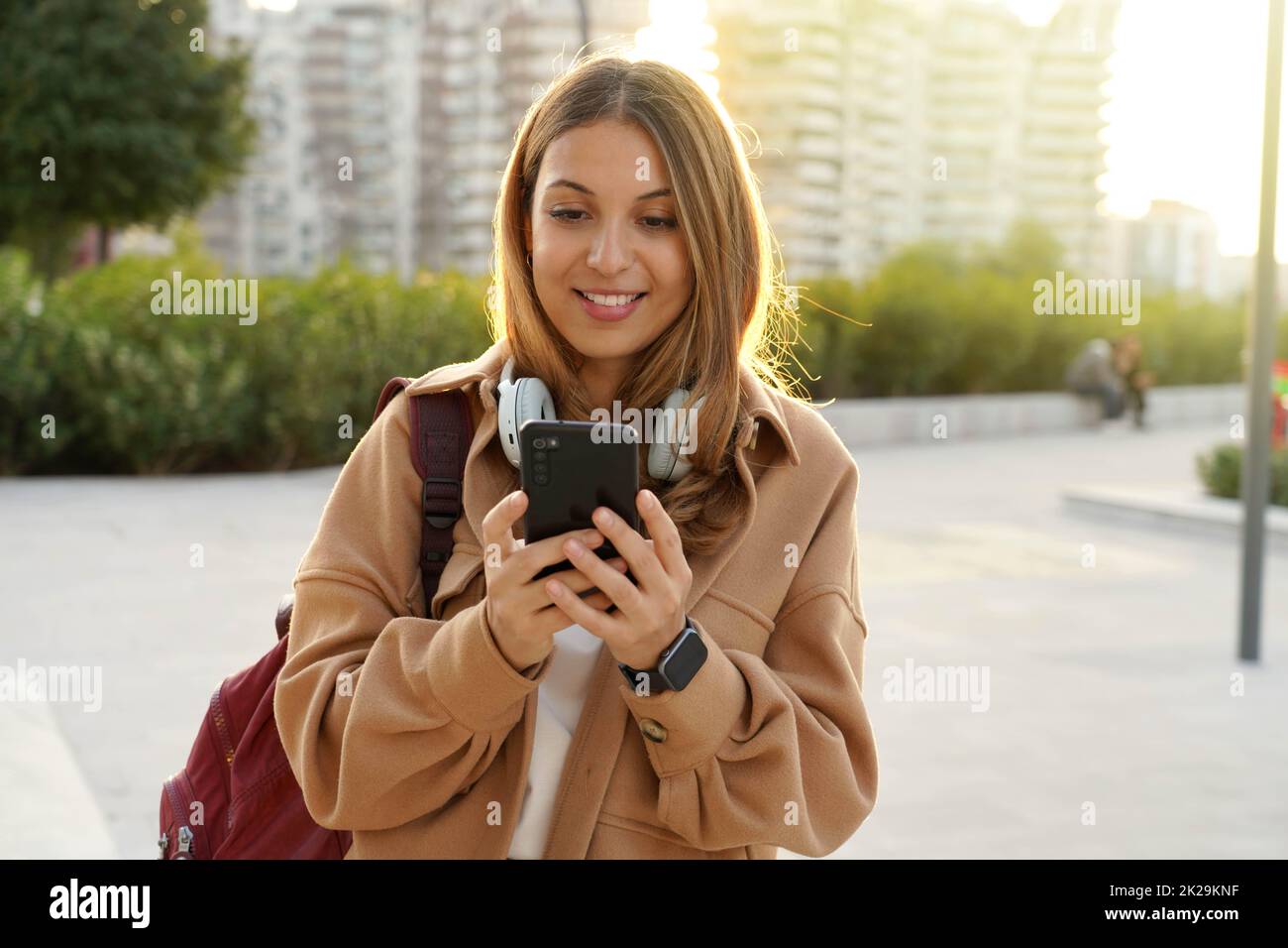 Sunshine Girl mit Mantel und Rucksack chattet bei Sonnenuntergang mit Smartphone-App auf der modernen Straße der Stadt. Nachhaltiges Leben. Generation der Millennials. Kopfhörermusik. Digital nativ. Intelligente Stadt. Stockfoto