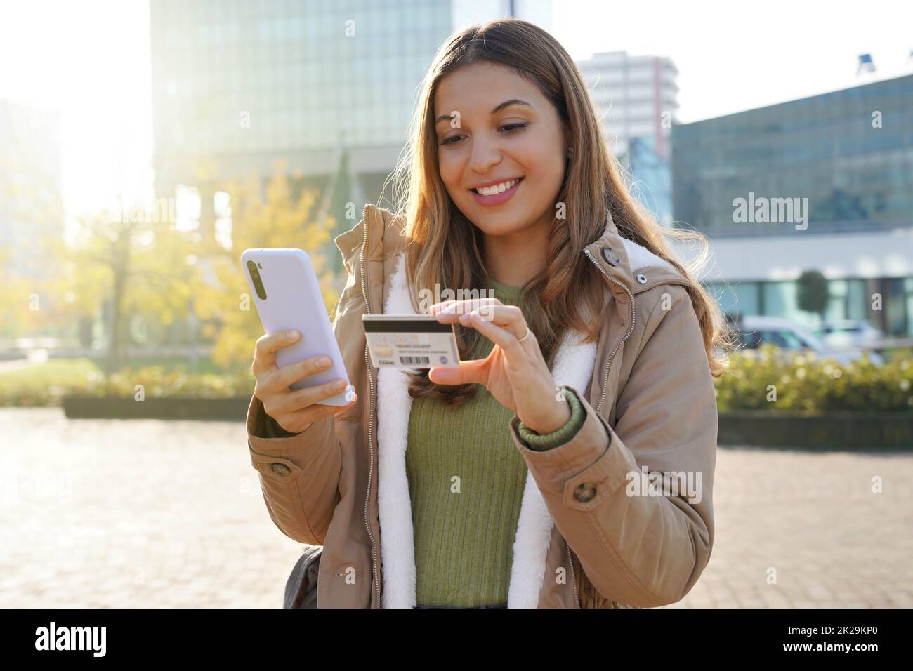 Portrait einer jungen Frau, die die Nummer der Kreditkarte auf dem Mobiltelefon eingibt, macht den Online-Kauf auf der Straße der Stadt einfach Stockfoto
