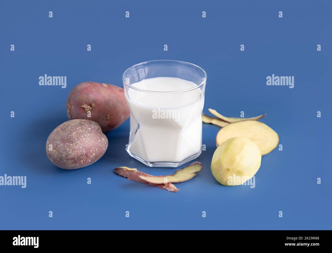 Ein Glas Gemüsekartoffelmilch auf einem blauen Tisch in der Nähe von Kartoffelknollen Stockfoto