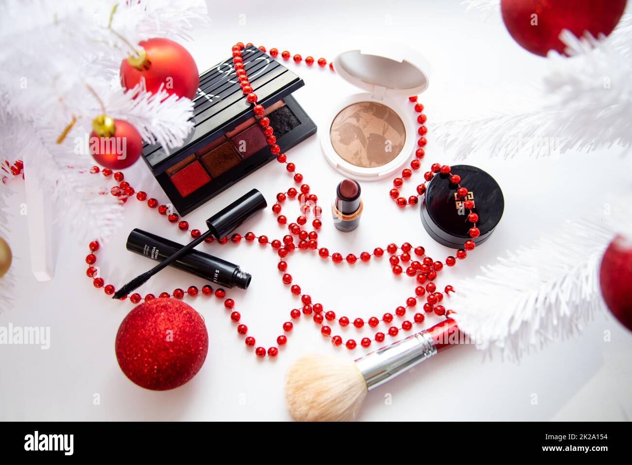 Puder und Bronzer, Lidschatten, Lippenstift, Rouge und ein Pinsel verschiedener Marken liegen auf weißem Hintergrund umgeben von weißen flauschigen Zweigen eines Weihnachtsbaums und roten Weihnachtsbällen. Stockfoto