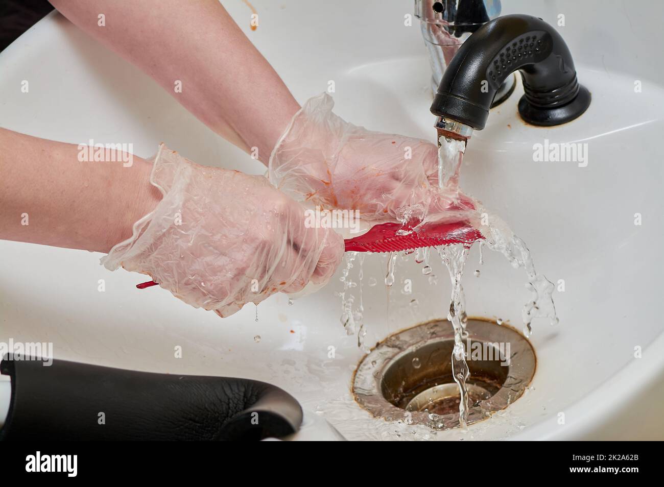 Frauenhände in Haarfärbehandschuhen waschen einen Kamm unter einem Wasserstrahl in einem weißen Waschbecken Stockfoto