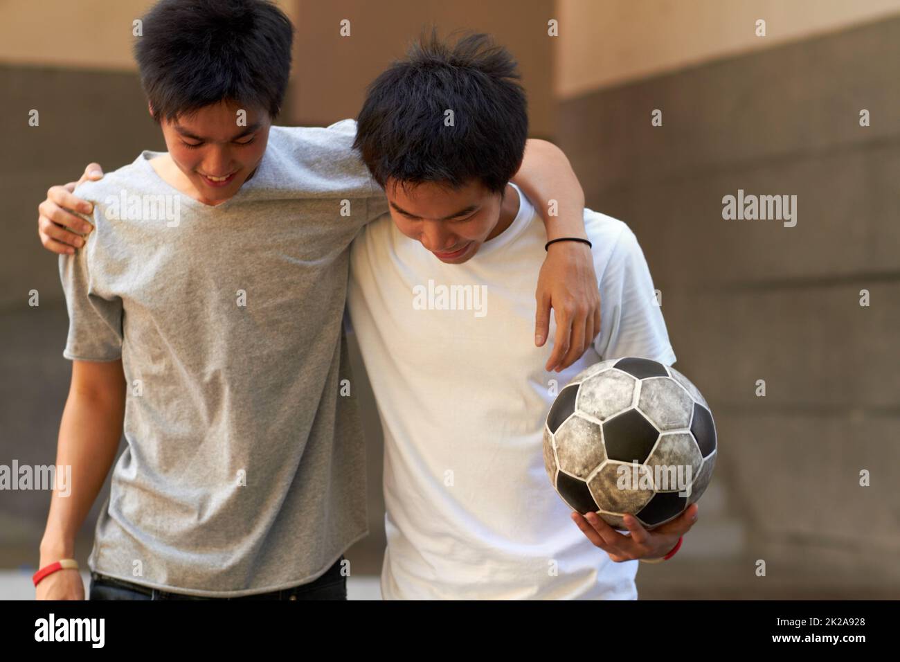 Gutes Spiel. Zwei asiatische Jungen, die mit ihren Armen umeinander gehen und einen Fußball halten. Stockfoto