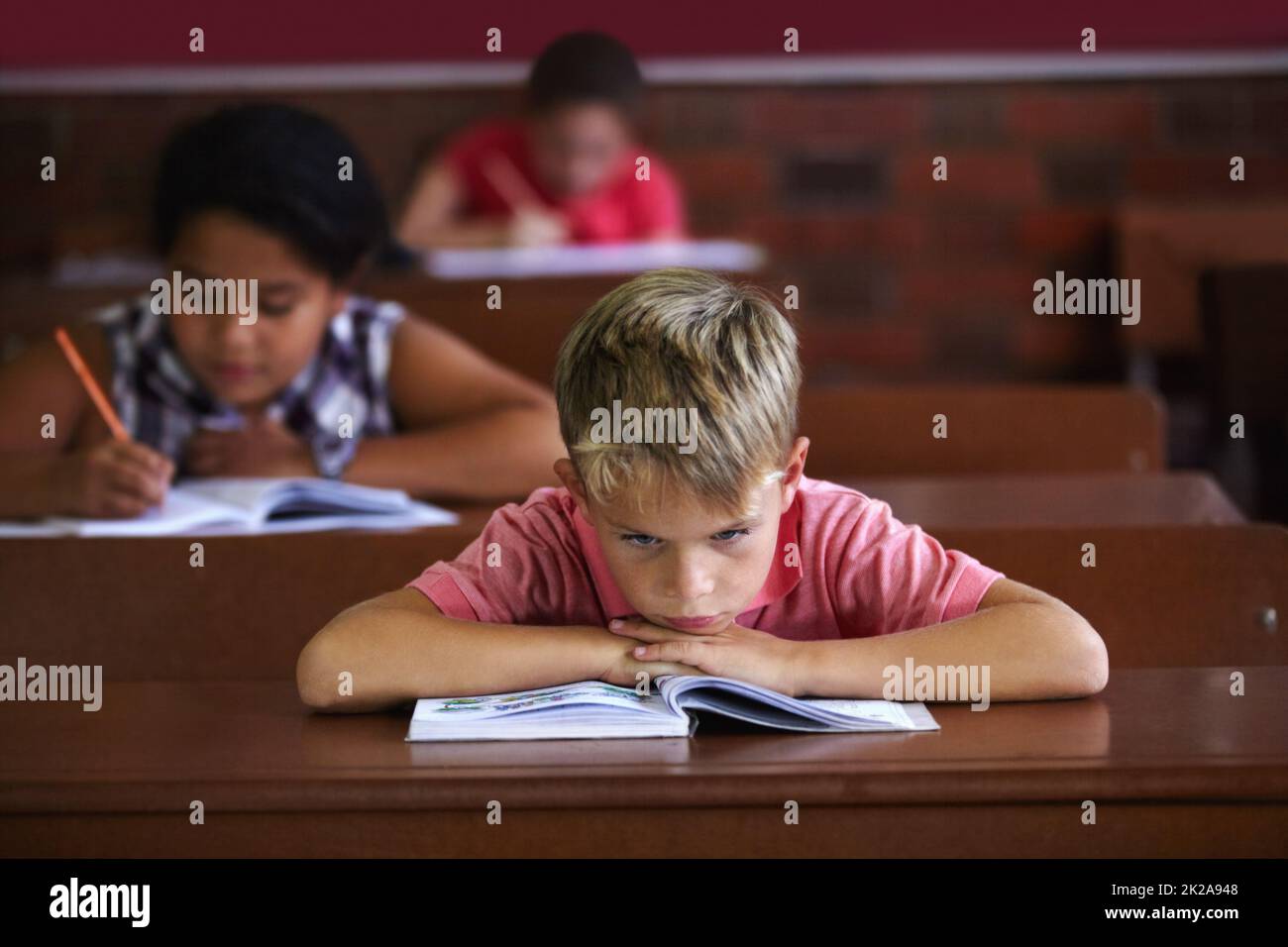 Die Schule ist sauer. Ein kleiner Junge, der seinen Kopf auf seinen Armen ruht, während er in einem Klassenzimmer sitzt und langweilig aussieht. Stockfoto