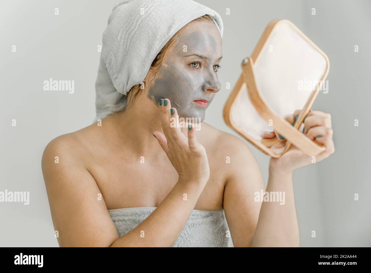 Die junge Frau legt graue kosmetische Tonmaske auf das Gesicht, während sie in den Spiegel schaut, ihr Haar und ihren Körper in ein Handtuch gewickelt, Seitenansicht. Nahaufnahme Stockfoto