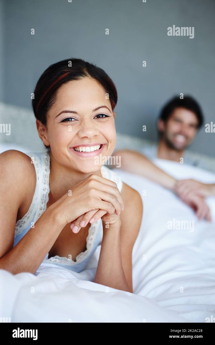 Ich liebe es, darin zu liegen. Porträt einer lächelnden Frau, die auf ihrem Bett liegt, mit ihrem Freund im Hintergrund. Stockfoto