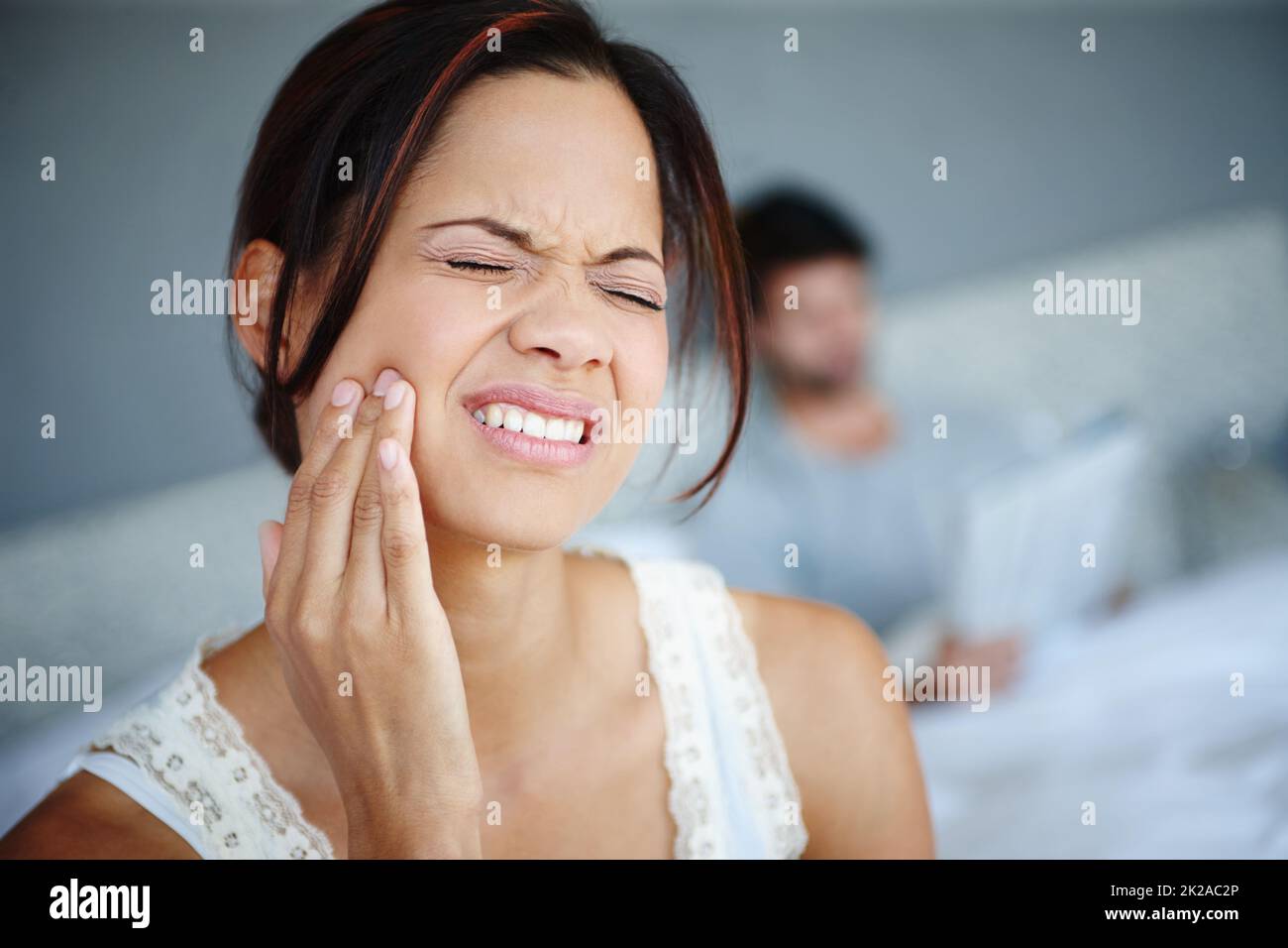 Zeit zum Zahnarzt zu gehen. Aufnahme einer Frau, die mit schweren Zahnschmerzen auf der Seite ihres Bettes sitzt und ihren Freund im Hintergrund hat. Stockfoto
