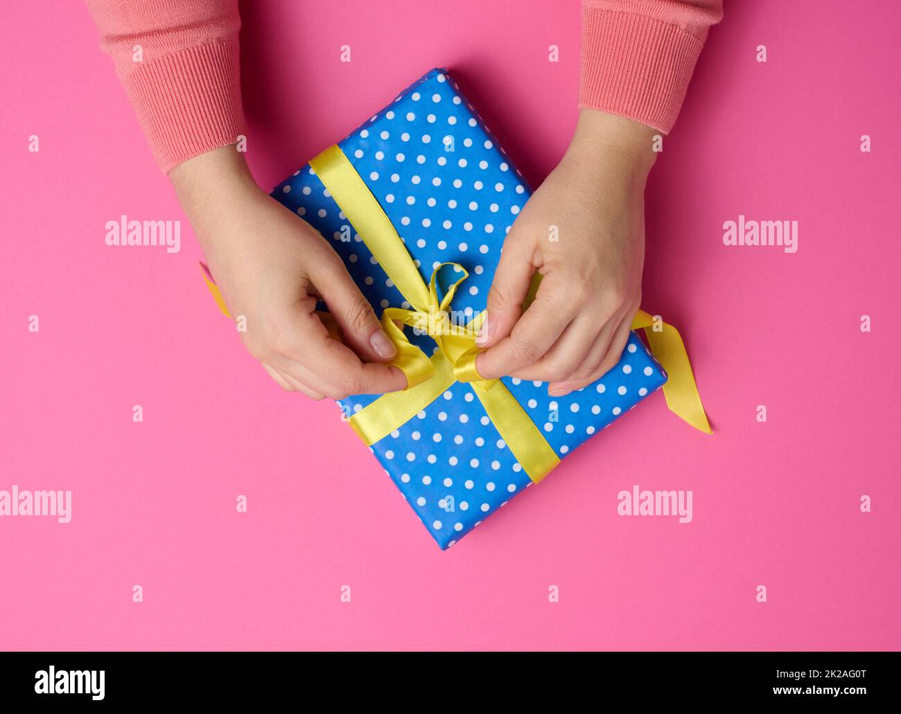 Weibliche Hand bindet ein Seidenband auf einer Geschenkbox, rosa Hintergrund. Vorbereitung auf den Urlaub Stockfoto