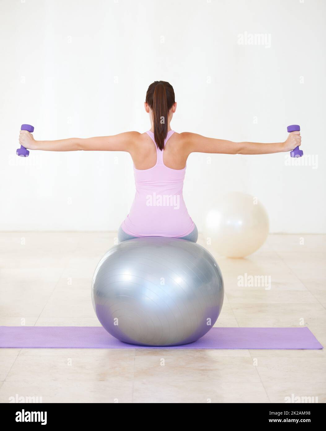 Die beiden Kernkomponenten ihres Workouts. Rückansicht einer jungen Frau, die auf einem Sportball sitzt und ein paar Kraftübungen mit Kurzhanteln macht. Stockfoto