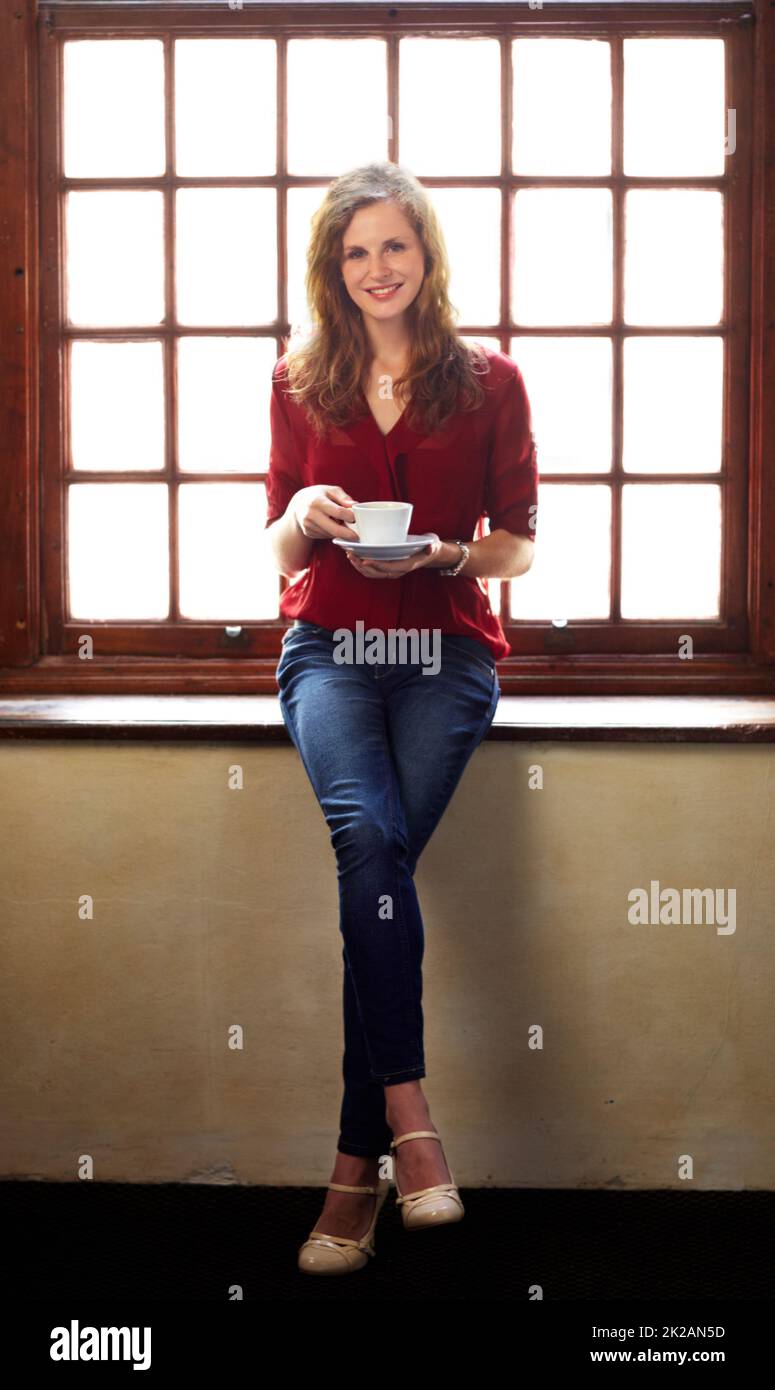 Den Tag richtig beginnen. Lächelnde junge Frau, die auf einer Fensterbank thront und eine Tasse Kaffee genießt - Porträt. Stockfoto