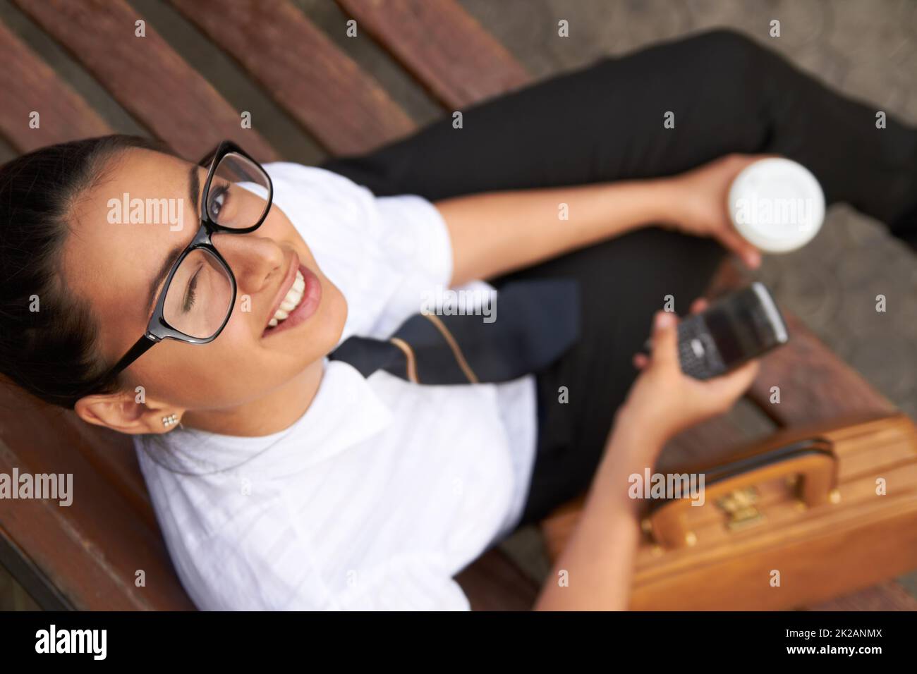 Ich lerne, was eure Supermacht ist. Eine schöne junge Frau winkelt an der Kamera, während sie auf einer Bank mit ihrem Handy, Kaffee und Tasche sitzt. Stockfoto