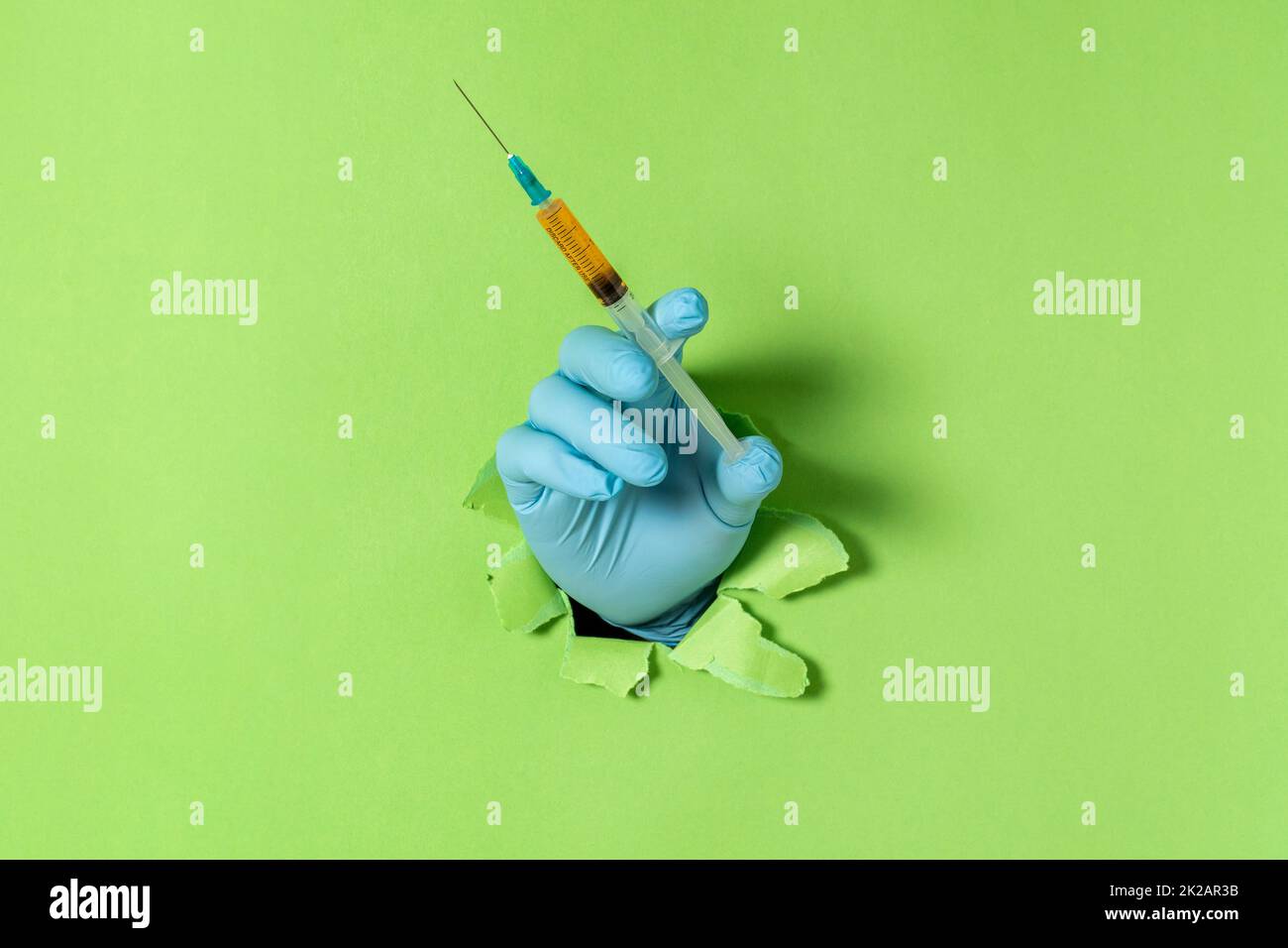 Hand in blauem Handschuh, der die Spritze durch ein zerrissenes grünes Papier hält Stockfoto