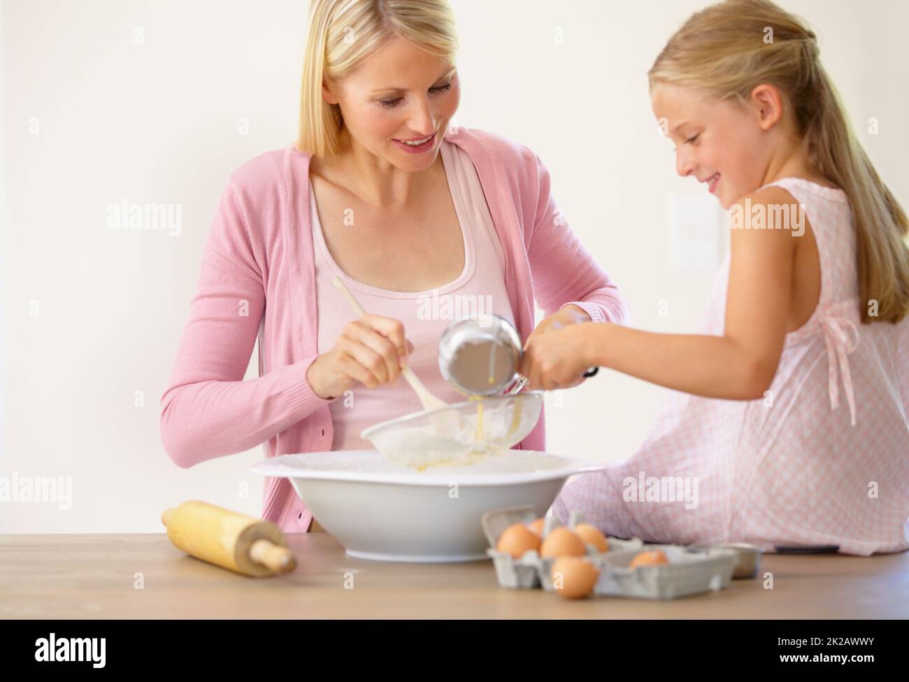 Misch alles in mit Mama. Ein kleines Mädchen, das auf einer Küchentheke sitzt und ihrer Mutter beim Backen hilft. Stockfoto