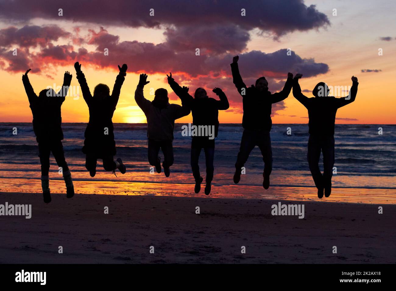 Erleben Sie die Awesomeness der Natur. Silhouette einer Gruppe von Menschen, die bei Sonnenuntergang am Strand springen. Stockfoto