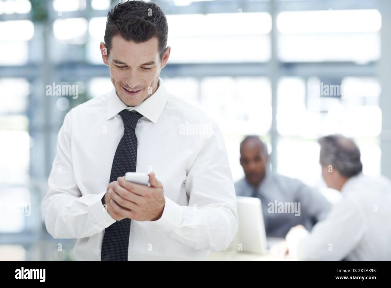 Eine sms an seine Familie senden. Ein junger Geschäftsmann schreibt vor zwei Kollegen auf seinem Handy. Stockfoto