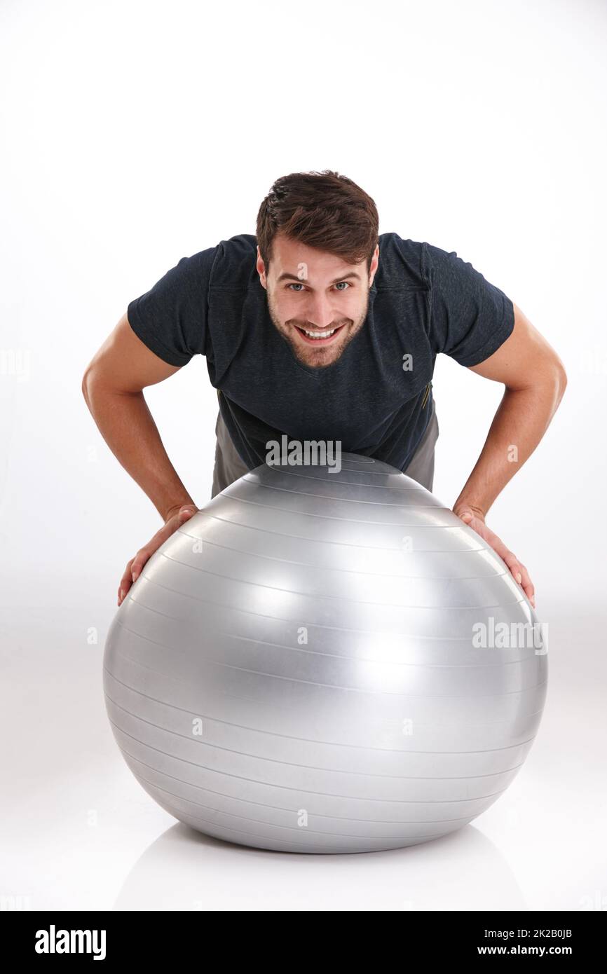 Konzentration auf ausgewogene Fitness. Studioaufnahme eines lächelnden jungen Mannes, der Liegestütze auf einem Übungsball macht. Stockfoto