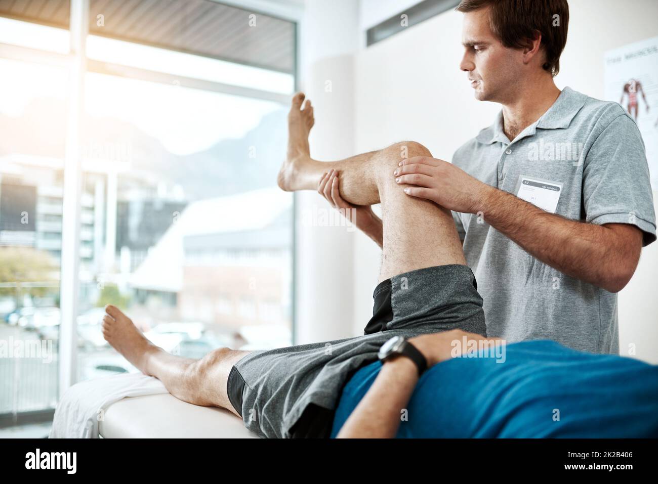 Ich werde Euch in kürzester Zeit reparieren. Aufnahme eines jungen männlichen Physiotherapeuten, der einem Klienten mit Beinübungen hilft, die auf einem Bett liegen. Stockfoto