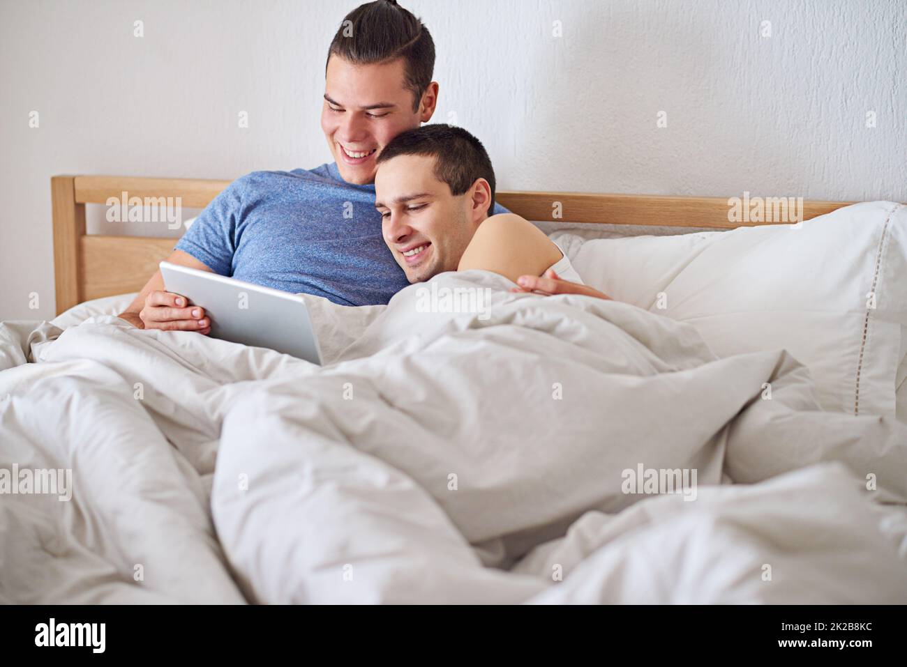 Ein junges schwules Paar, das ein digitales Tablet benutzt, während es sich im Bett entspannt. Stockfoto