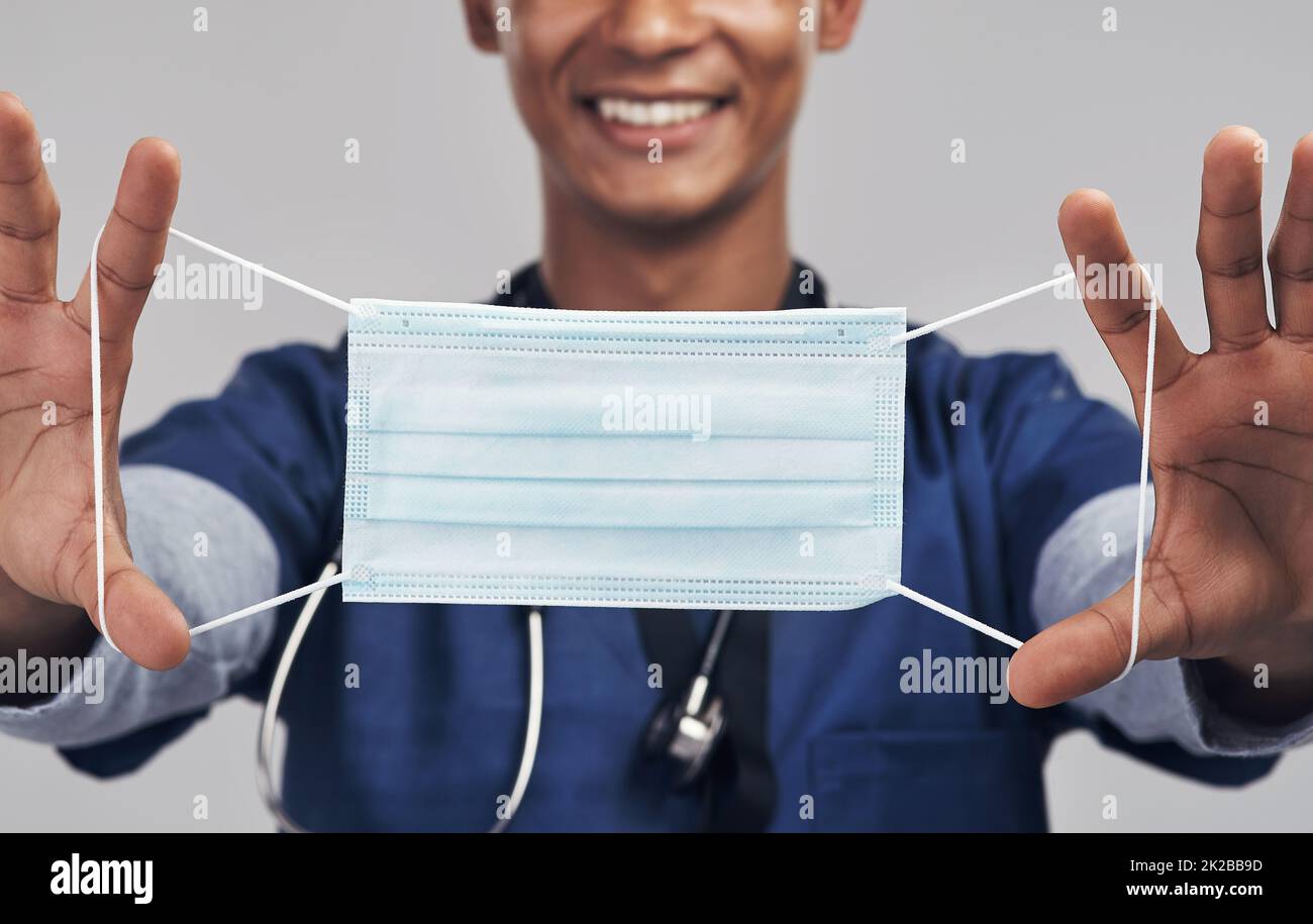 Trage deine Maske. Aufnahme eines Krankenpflegers, der eine OP-Maske hochhält, während er vor einem grauen Hintergrund steht. Stockfoto